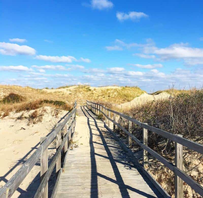 Wooden boardwalk leading through sand dunes under a blue sky with scattered clouds.