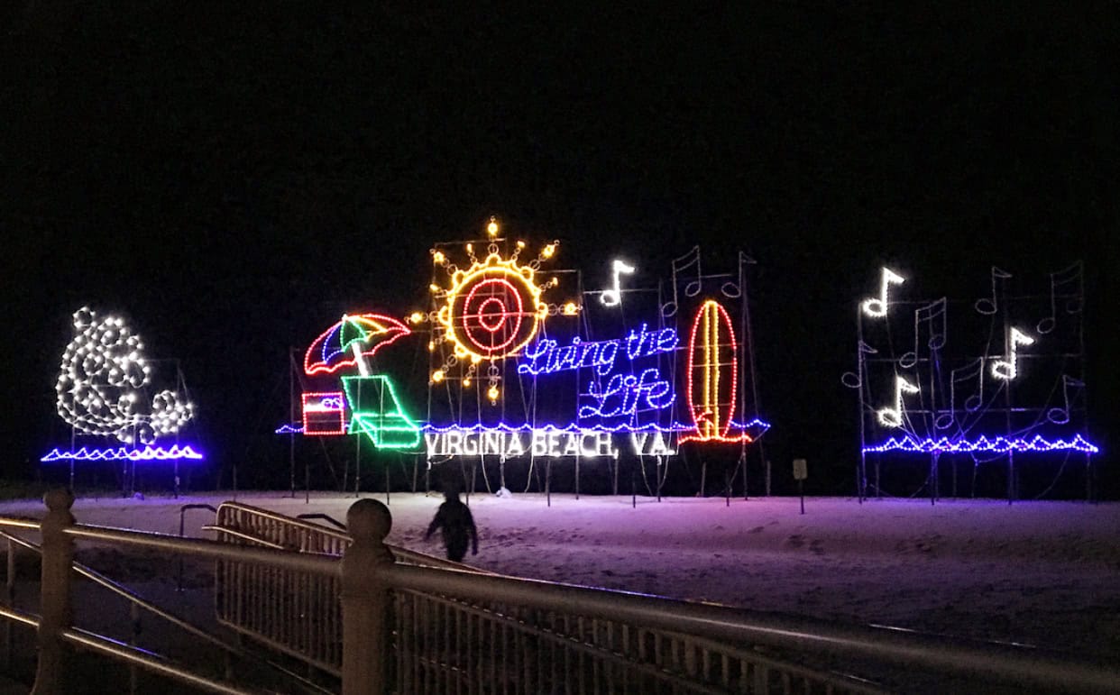 A colorful light display on the oceanfront features a tree, beach chair, umbrella, wave, sun, surfboard, and musical notes with the text "Living the Life Virginia Beach, VA" at night.