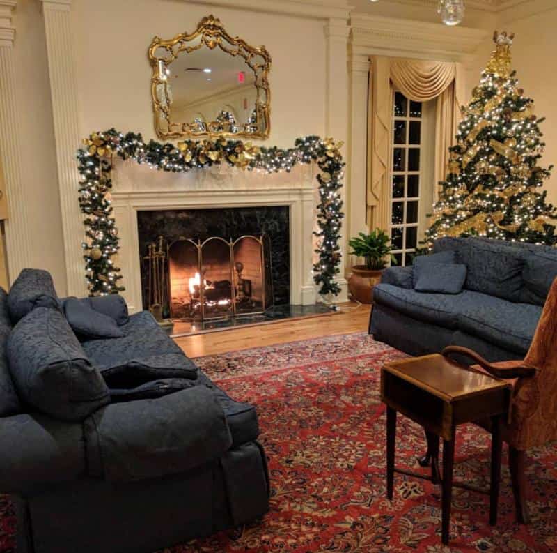 A cozy living room with a lit fireplace, two blue sofas, a decorated Christmas tree, and a garland on the mantel. An ornate mirror hangs above the fireplace.