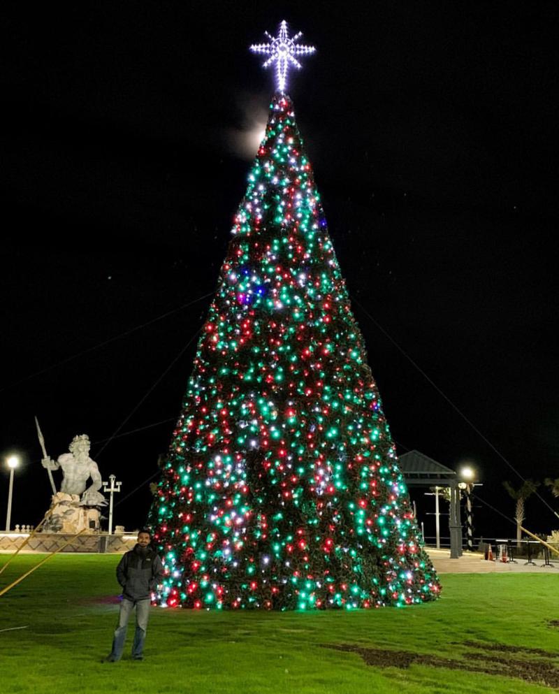 A person stands in front of a large Christmas tree adorned with colorful lights and topped with a glowing star, set against a nighttime backdrop.