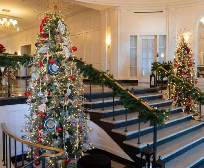 Elegant hotel lobby with grand staircase, decorated for the holidays with ornate Christmas trees, garlands, and festive lights.