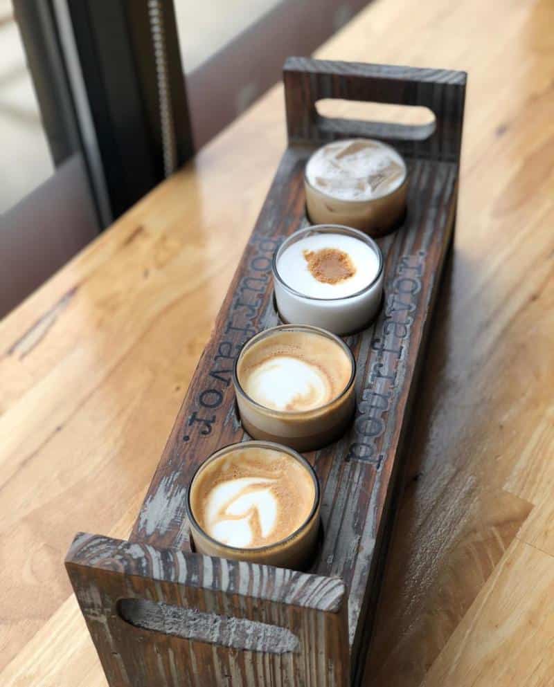 A wooden tray holds four cups of coffee with different latte art, placed on a wooden table near a window.