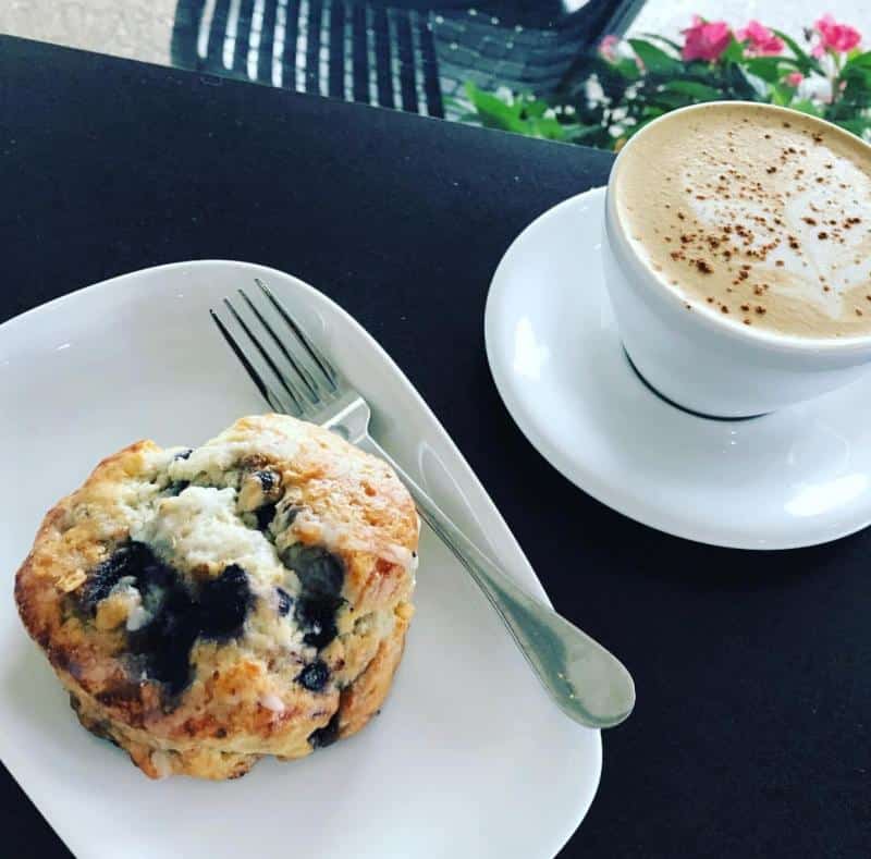 A blueberry scone on a white plate with a fork next to a cup of cappuccino on a black tabletop.