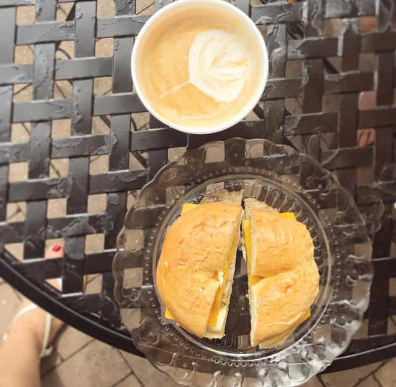 A cup of coffee with latte art sits on a black metal table next to a clear plate holding a cut bagel sandwich with cheese.