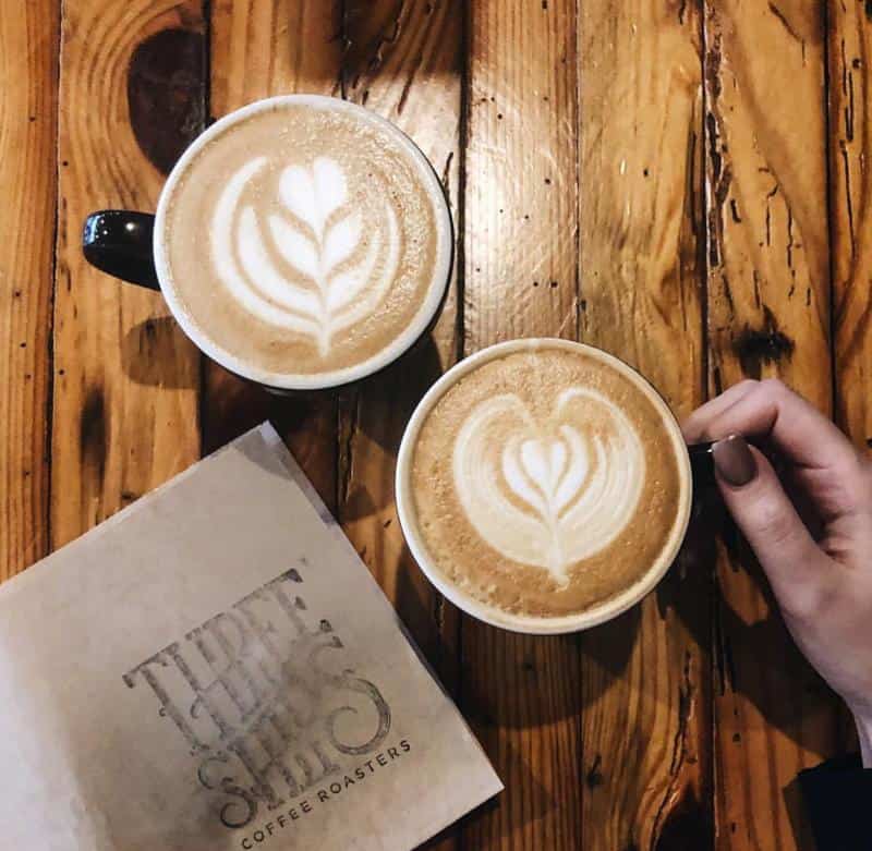 Two cups of latte with leaf latte art on a wooden table. A hand holds one cup. A Three Ships Coffee Roasters menu is visible.