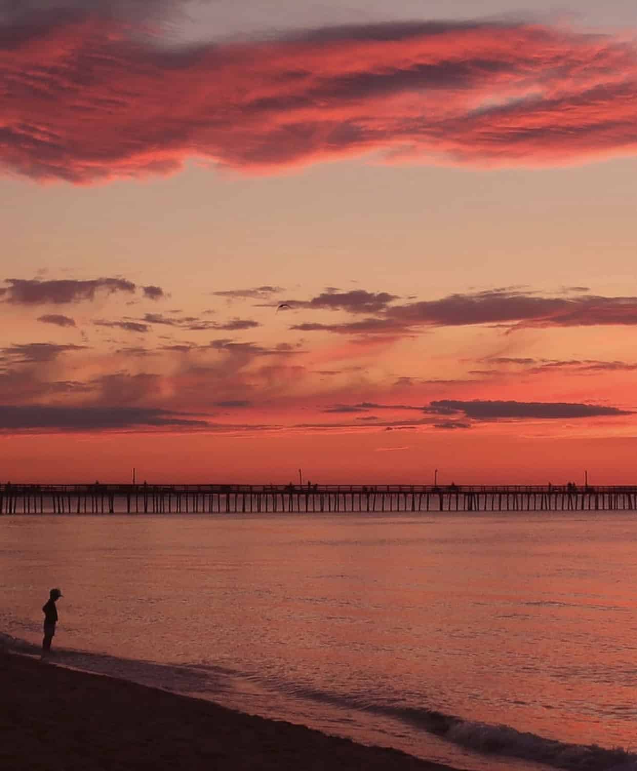 A person stands by the shoreline at sunset, with pink and orange clouds above and a pier stretching across the horizon.