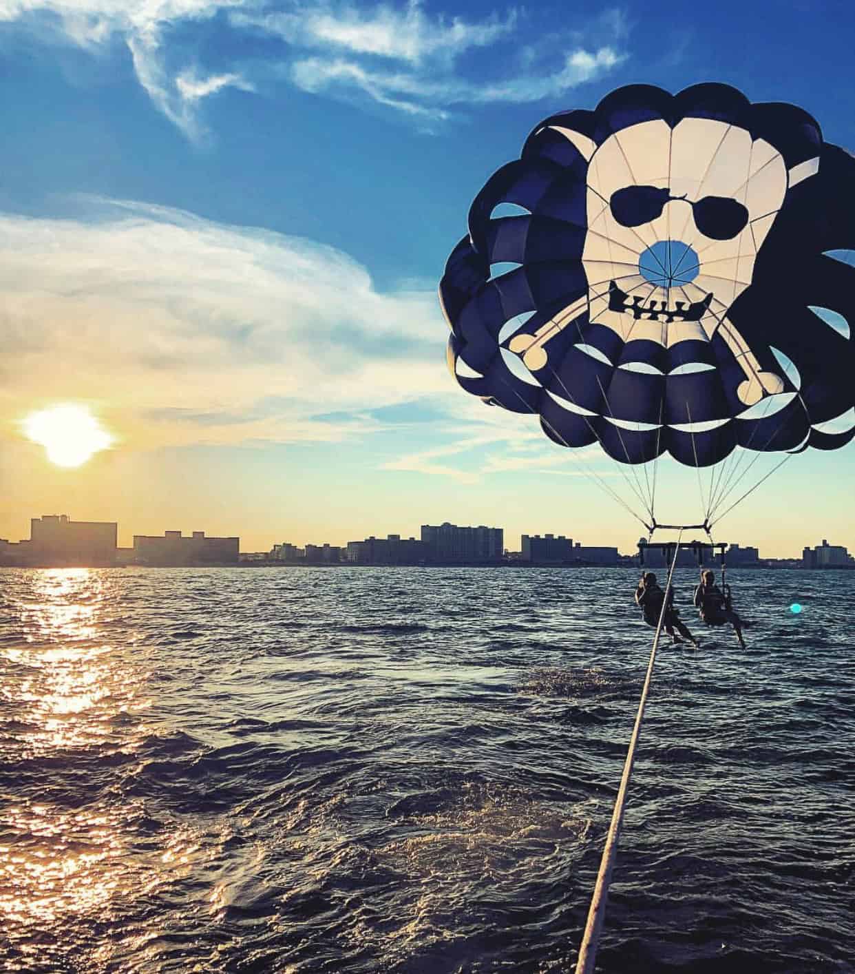 Two people parasailing over the ocean with a parachute featuring a skull and crossbones design, silhouetted against a sunset and city skyline.