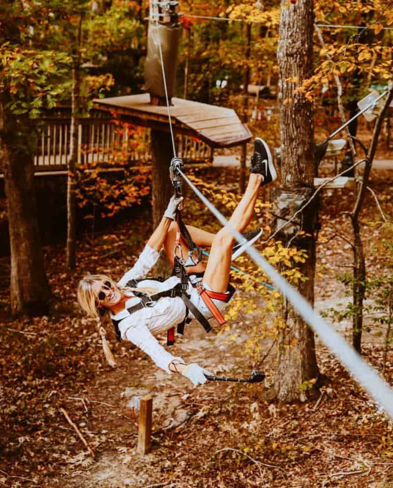 A person wearing a harness is upside down on a zip line in a forested area with autumn leaves visible.