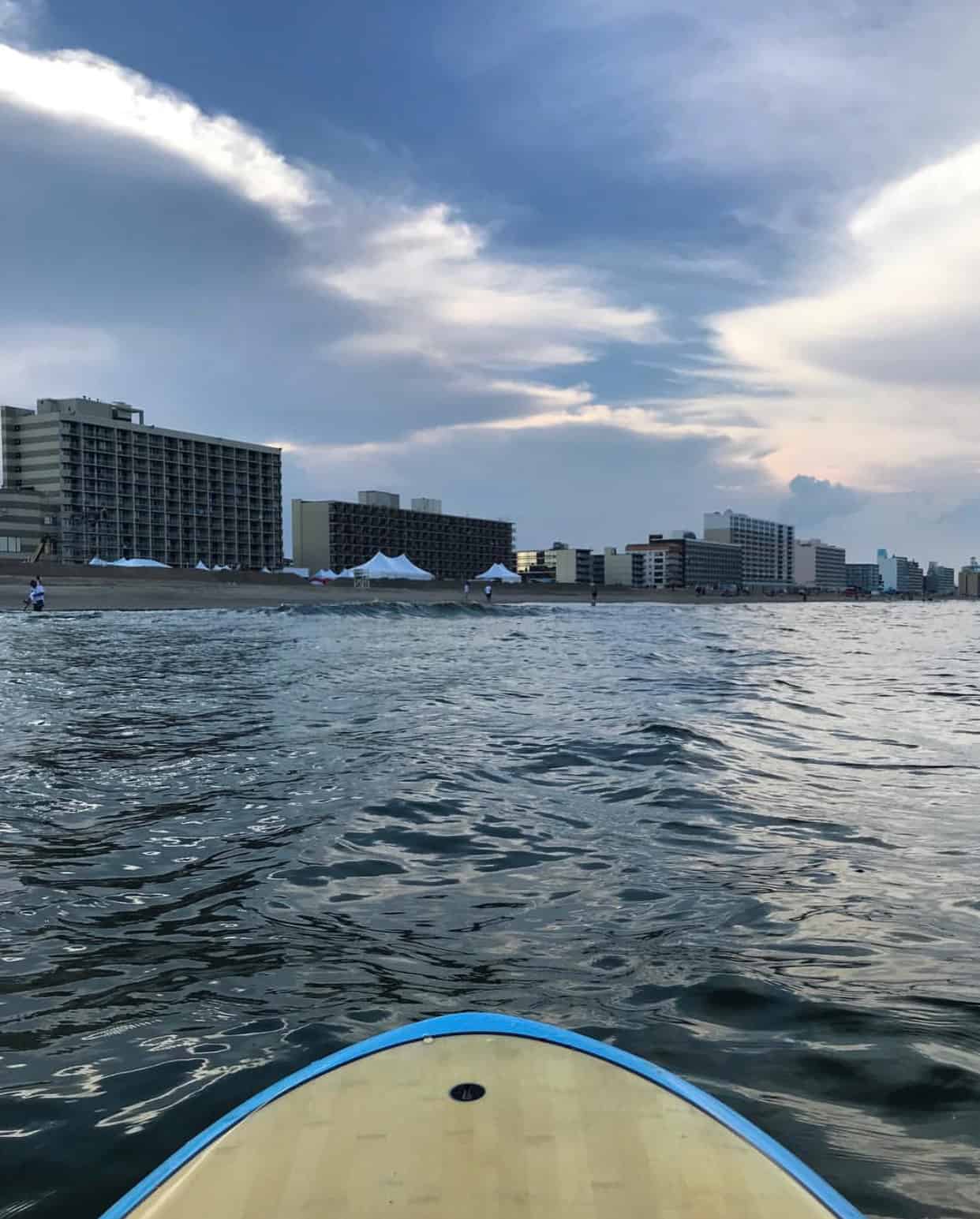 View from a paddleboard on the water, facing a shoreline with tall buildings under a cloudy sky.