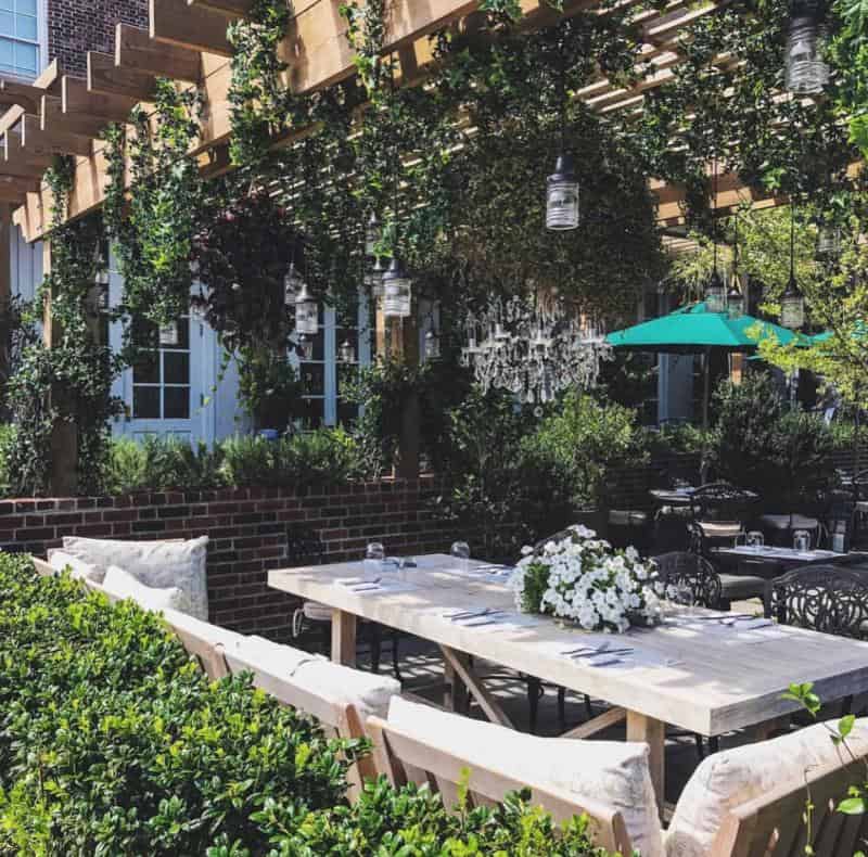 Outdoor patio with a wooden pergola covered in greenery, hanging lights, a table set with flowers, and cushioned chairs. An umbrella is visible in the background.