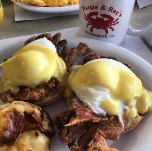 Breakfast featuring eggs Benedict with soft-shell crab on a plate, alongside a Margie & Ray's Sandbridge, VA mug.