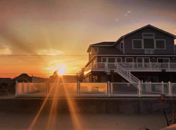 Sunset view of a two-story beach house with a large porch and white fence, located by the water.