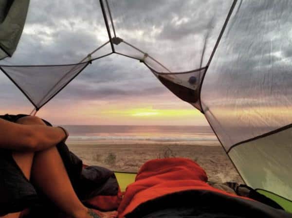View from inside a tent overlooking a beach at sunset, with two people sitting inside. The sky is cloudy with hues of orange and purple.