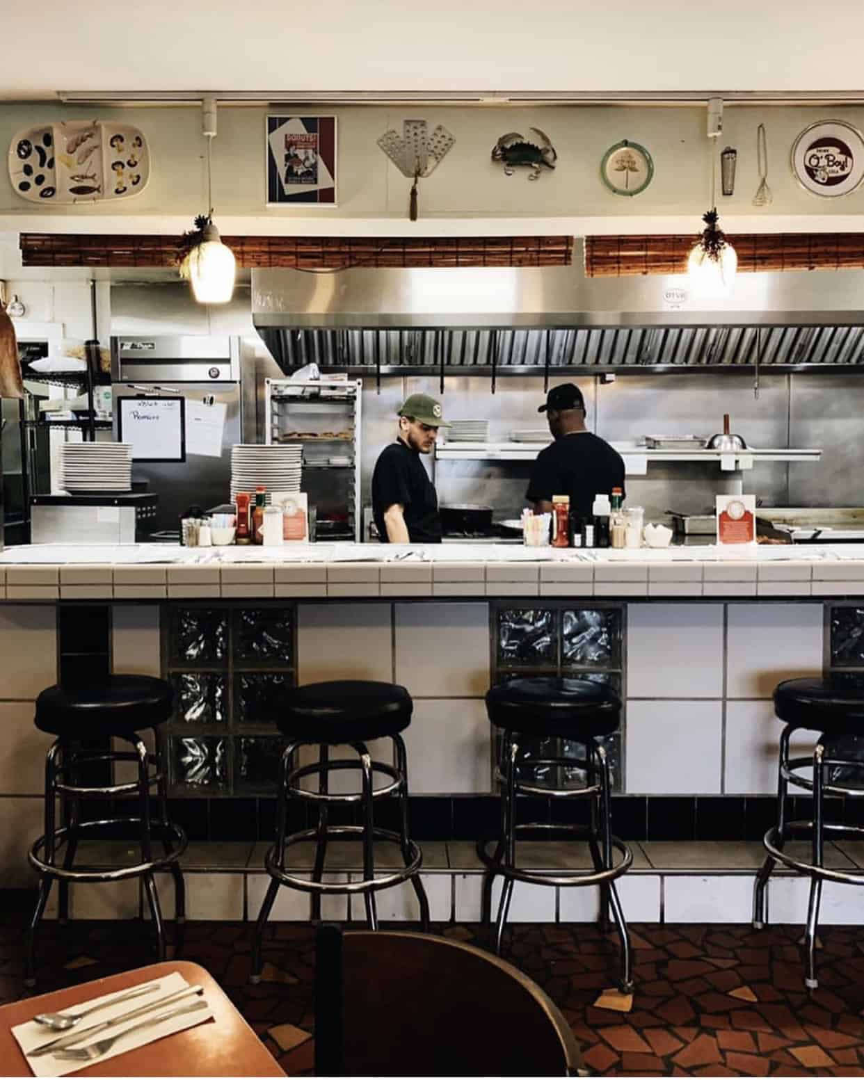 Two people work behind a retro-style diner counter with stools and light fixtures. Decor includes art, shelves, and kitchen equipment in the background.