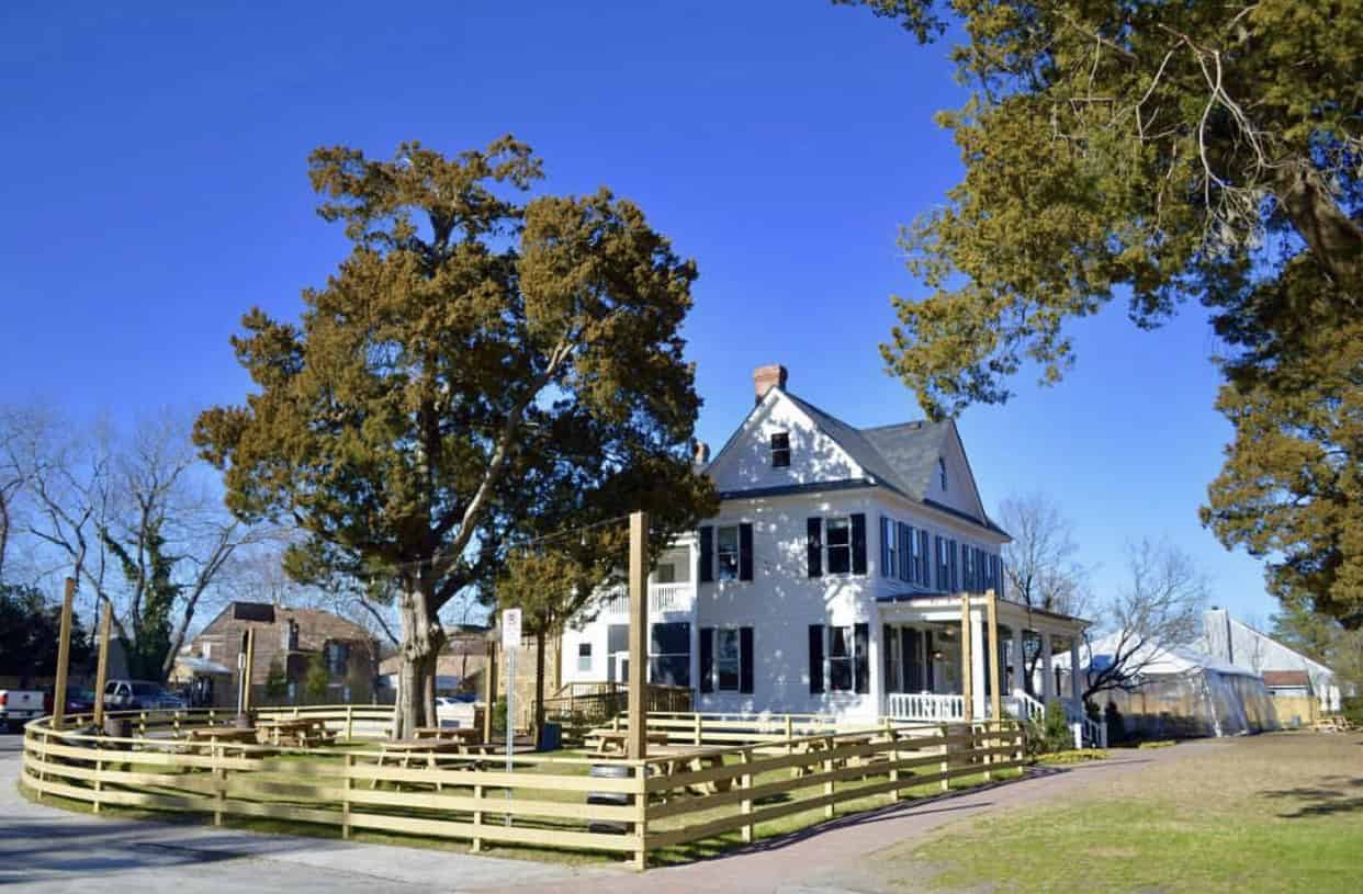Large white house with dark shutters, surrounded by a wooden fence and trees under a clear blue sky.