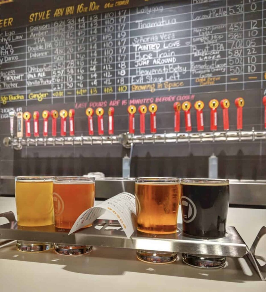 At a Virginia Beach brewery, a flight of four beers in glasses sit on a tray with a tasting menu, in front of a wall displaying a variety of draft options and their details.