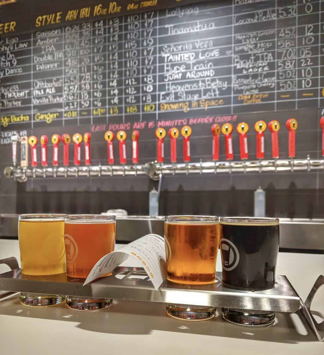 A flight of four beers in glasses on a tray with a tasting menu, in front of a wall displaying a variety of draft options and their details.