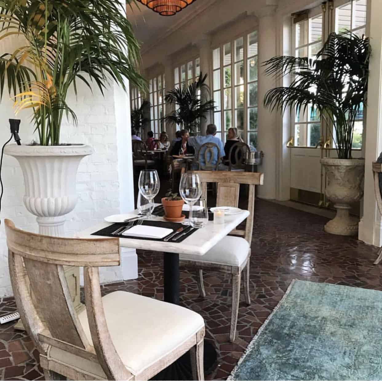 Elegant restaurant interior with a marble table set for two, surrounded by tall potted plants and wooden chairs on a tiled floor. Large windows line one wall, revealing several diners.