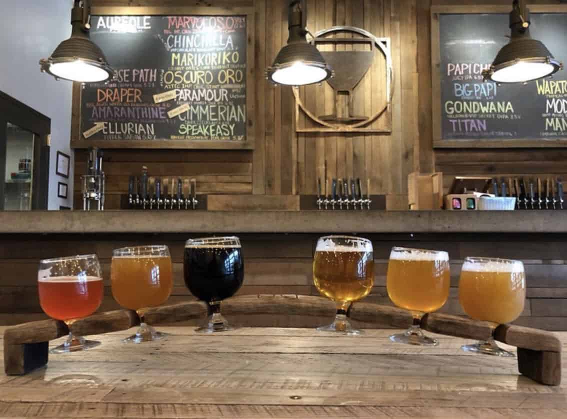 Six glasses of beer in varying colors are arranged on a wooden table in a rustic brewery setting with menu boards in the background.