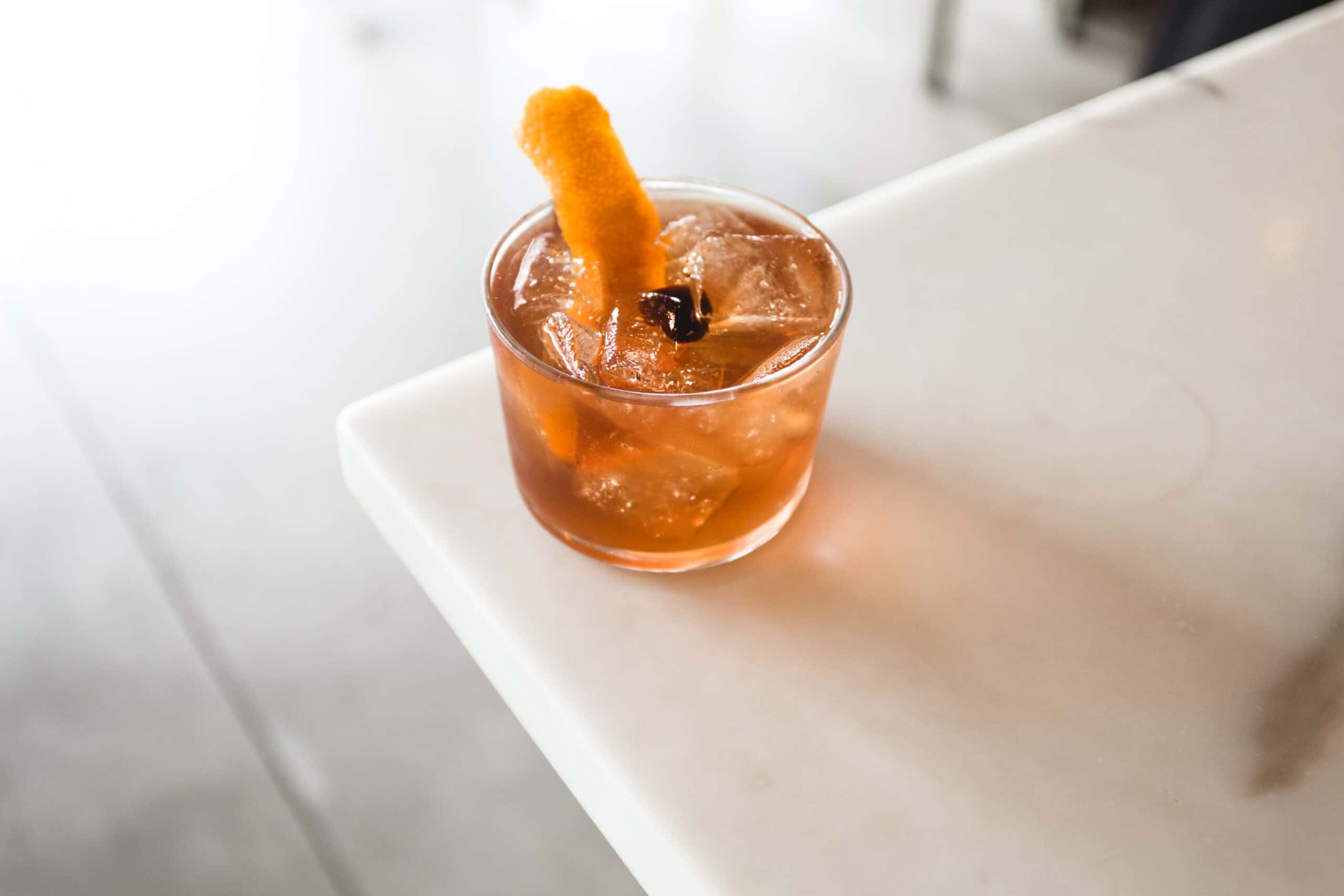 A cocktail with ice and an orange peel garnish sits on a white marble surface.