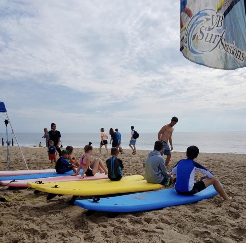 People sitting on surfboards on a sandy beach with the ocean in the background. A flagged sign reads "VB Surf Sessions.