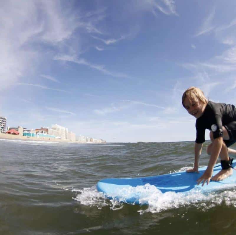 Child surfing on a blue surfboard in the ocean near a city shoreline, under a clear blue sky.