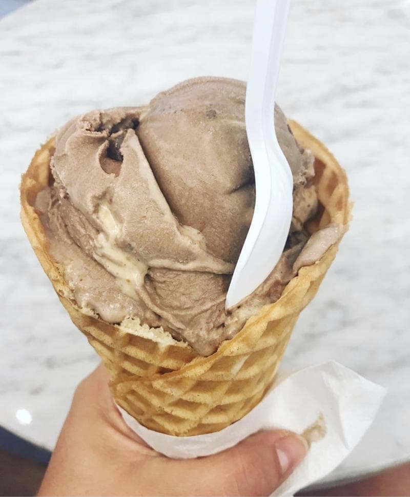 A hand holds a waffle cone with scoops of chocolate ice cream, a white plastic spoon is inserted, and a napkin wraps the cone.
