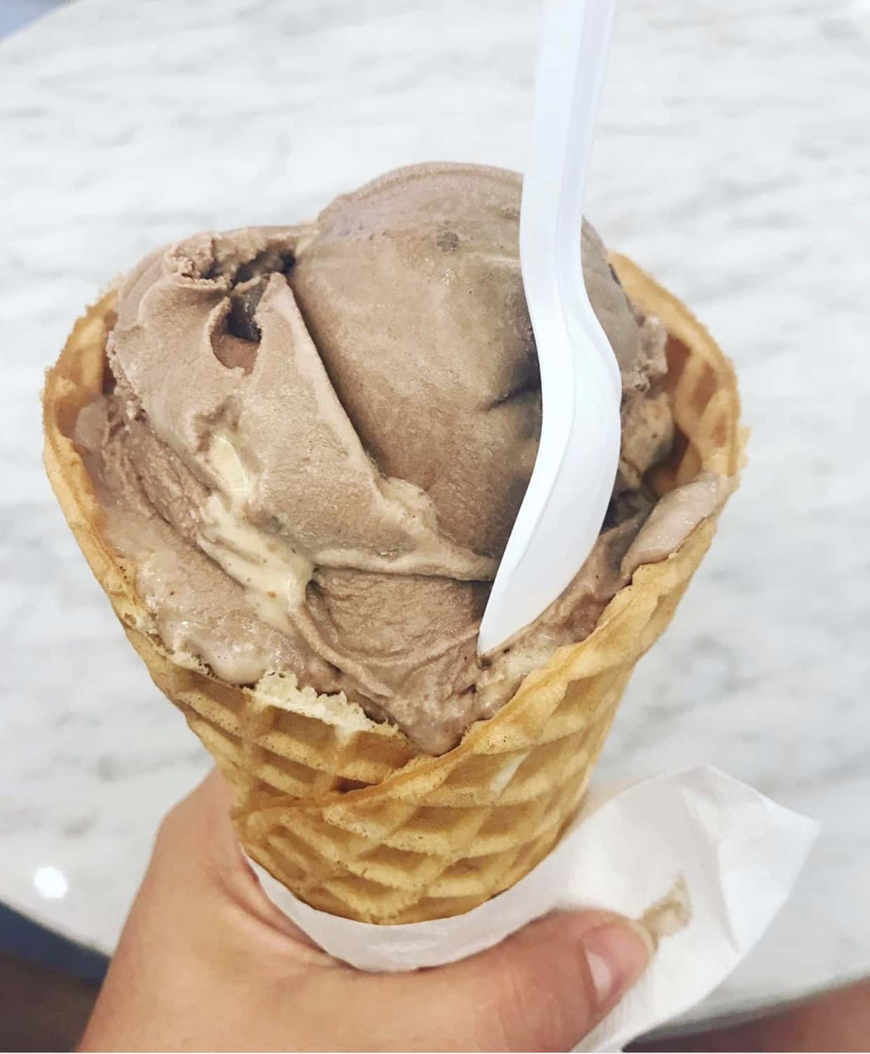 Hand holding a waffle cone with two scoops of chocolate ice cream and a plastic spoon, against a marble surface background.