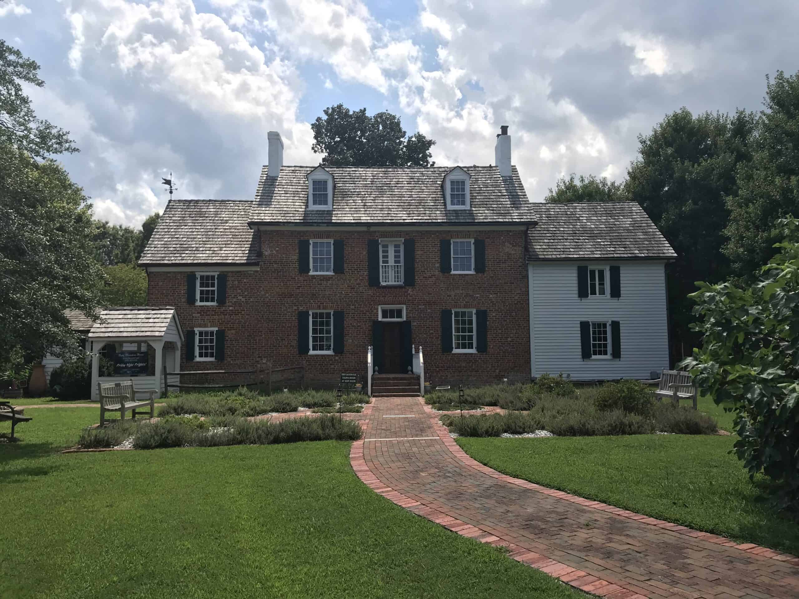 A two-story historic brick house with white siding on one section, surrounded by lush greenery and a red brick path, under a partly cloudy sky.