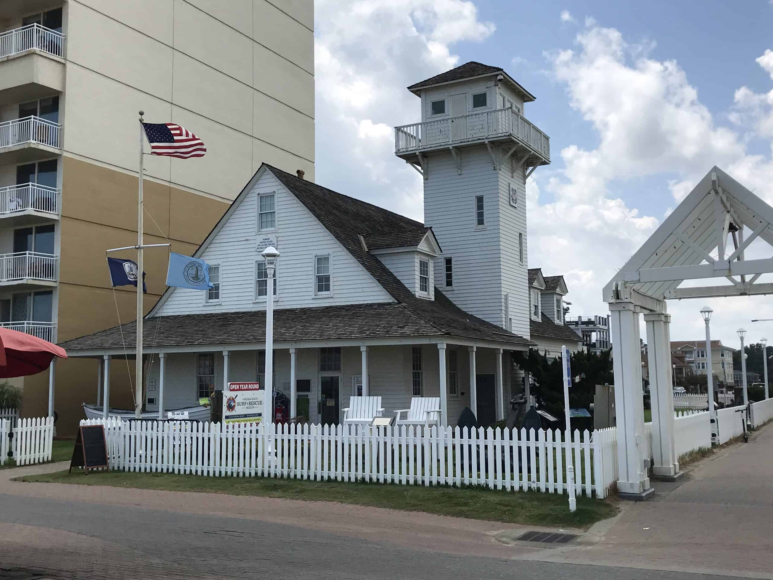 White wooden building with a tower and a picket fence, next to a high-rise. Flags are waving, and the sky is cloudy.