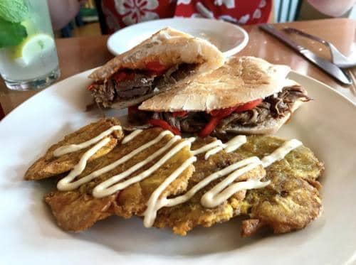 A sandwich with beef and red peppers is served on a white plate, accompanied by fried plantains drizzled with a creamy sauce. A glass with a lemon slice is in the background.