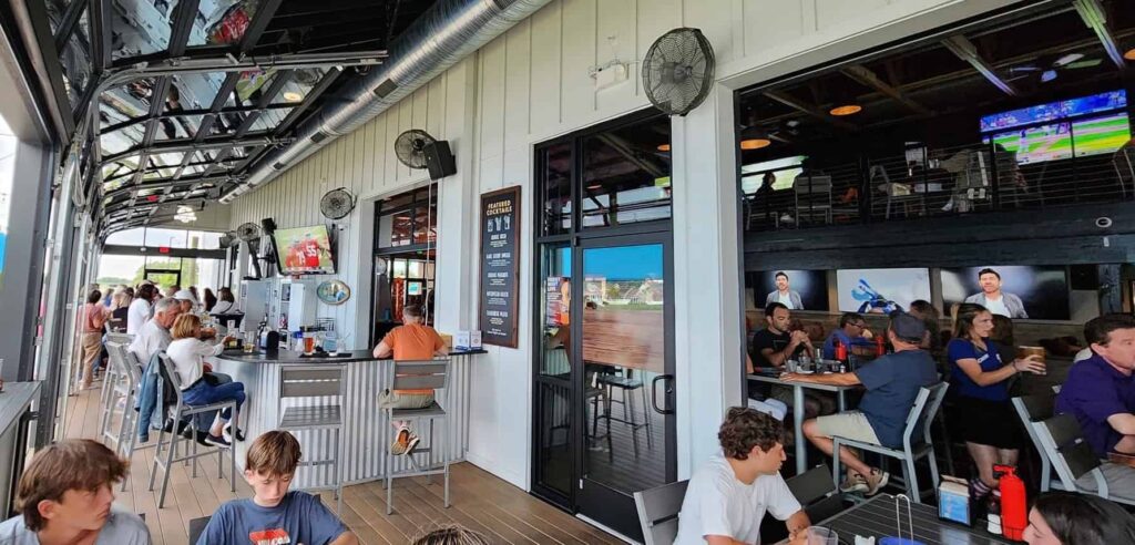 People dining and socializing at a sports bar with outdoor seating. Multiple TVs display sporting events. Fans are on the ceiling, and large windows provide views of the outside.