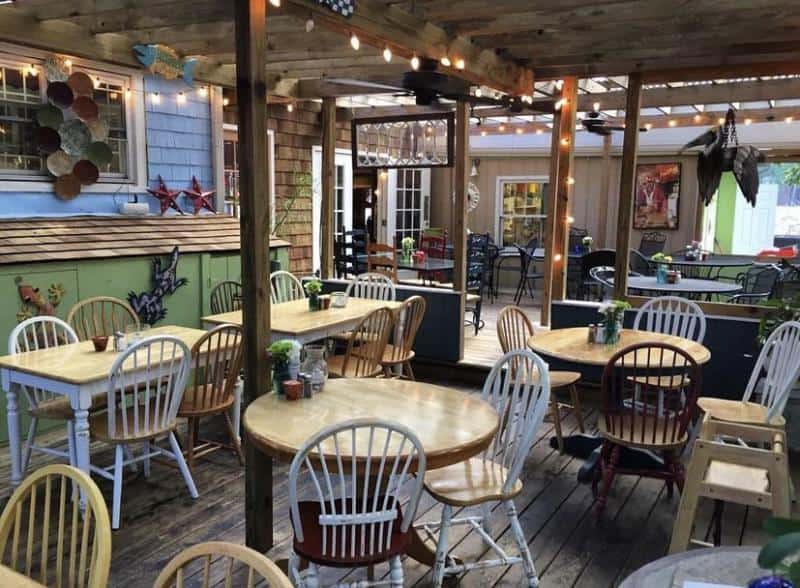 Outdoor café seating area with wooden tables and chairs, surrounded by rustic decor and string lights hanging overhead.