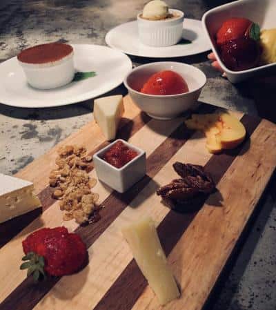 Cheese and fruit platter with nuts, jams, and dessert bowls on a wooden board and table.