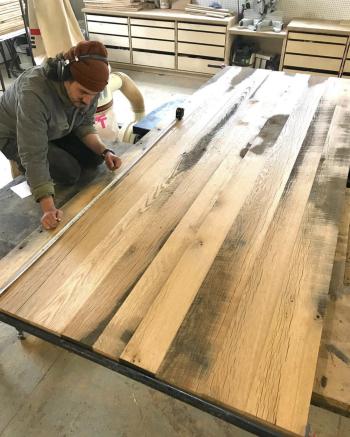 Person measuring a large wooden table in a workshop, wearing a beanie and headphones.