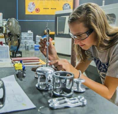 A person wearing safety goggles works on a metal component at a workstation surrounded by tools and parts.