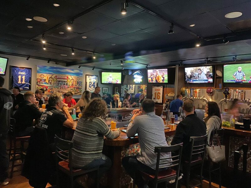 People are seated at a bar, watching various sports games on multiple screens. The walls are decorated with sports jerseys and paintings.