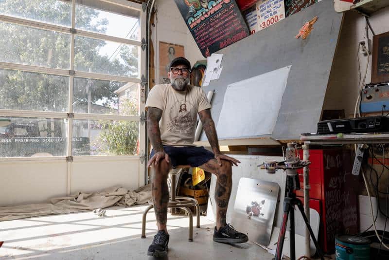 Bearded person with tattoos sits on a stool in an art studio, in front of a large easel, surrounded by paintings and posters. Sunlight enters through a window.