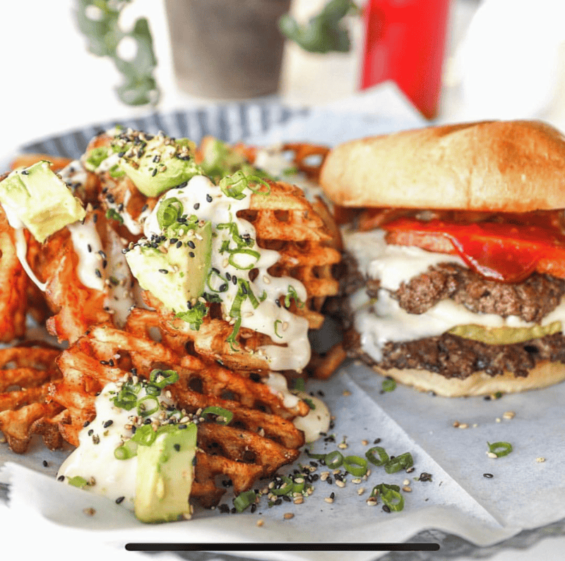 A burger with cheese, pickles, and red peppers is on a plate beside waffle fries topped with avocado, cheese sauce, and green onions.