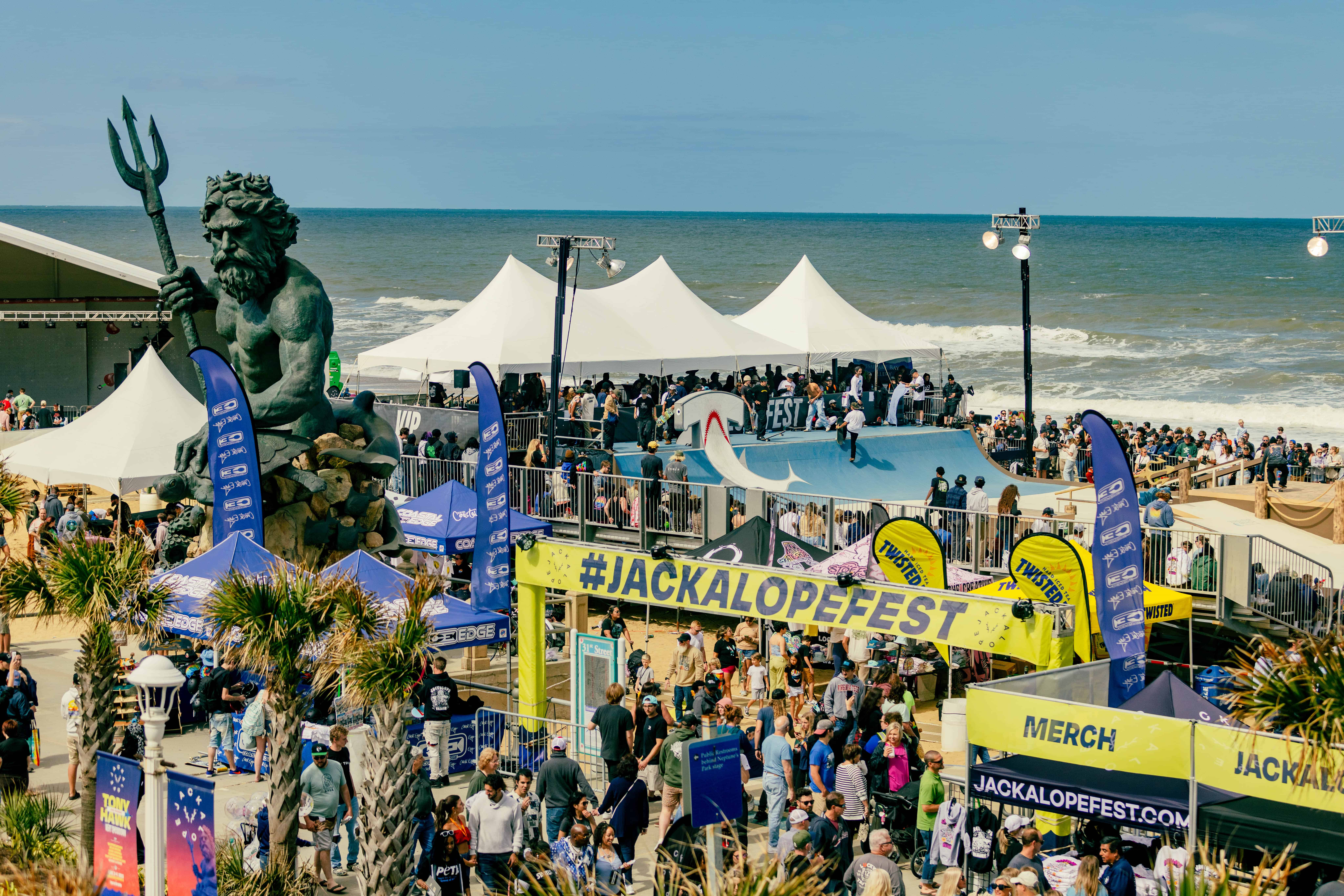 Crowd at an outdoor event with a stage and #JACKALOPEFEST signage. A large statue holding a trident overlooks the scene. Ocean waves are in the background.