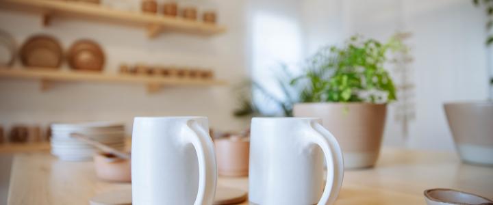 Two white mugs on a wooden table in a kitchen with shelves of pottery and a potted plant in the background.