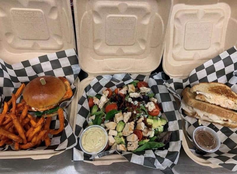 Three takeout containers: sweet potato fries with a burger, a salad with grilled chicken, avocado, tomatoes, a dressing cup, and a grilled sandwich with a sauce cup, all on checkered paper.