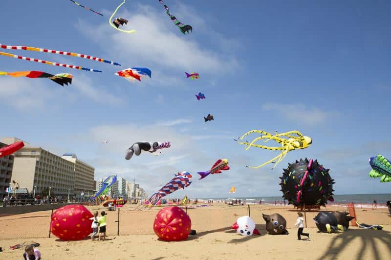 Colorful kites of various shapes and sizes flying above a sandy beach with buildings in the background on a clear day.