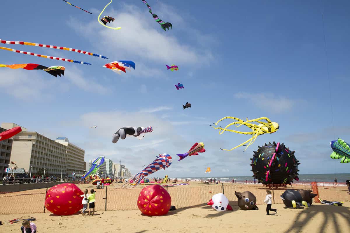 Colorful kites and inflatable structures float above Virginia Beach with several people, buildings, and a partially cloudy sky.