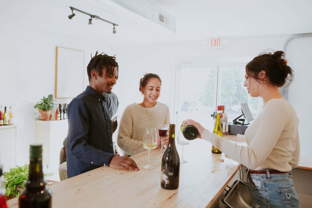 Three people at a wine bar: a server pours wine for two smiling patrons, one with a clear drink and one with a dark drink. Bottles and glasses are on the wooden counter.