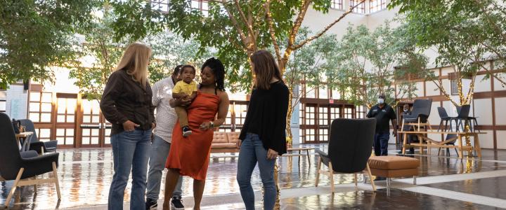 Four people converse in a spacious, indoor area with large trees and modern furniture. One person holds a small child.