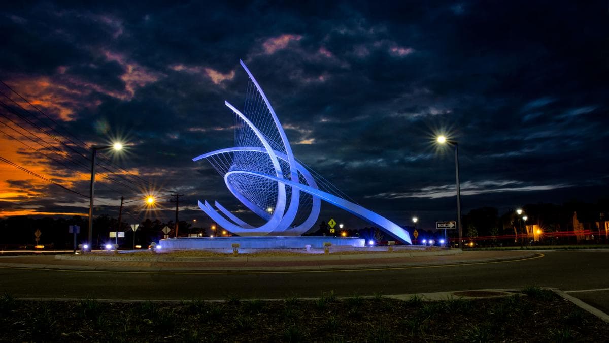 Illuminated abstract sculpture with blue lights at a roundabout, set against a dramatic evening sky with streetlights and a sunset.