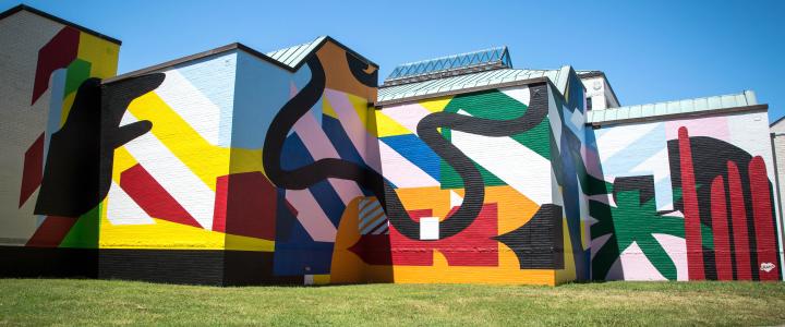 A vibrant mural with bold abstract shapes and patterns covers a building exterior under a clear blue sky.