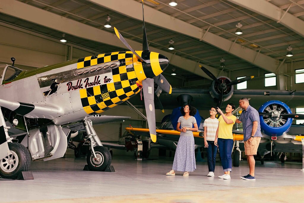 Four people stand in an aircraft hangar, observing a vintage propeller plane with a yellow and black checkerboard pattern on its nose. Other airplanes are visible in the background.