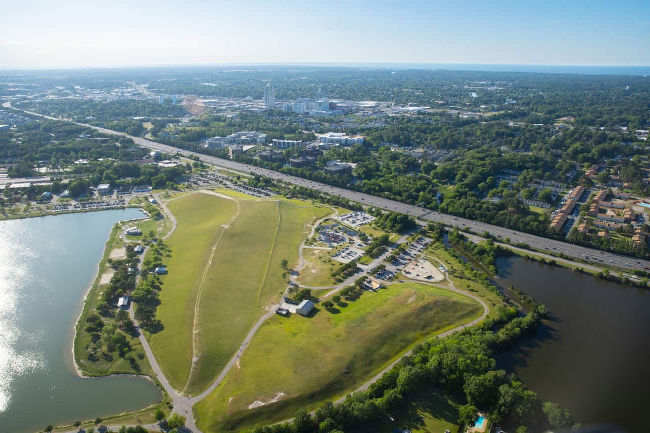 Aerial view of a green park bordered by a lake, surrounded by urban development and highways under a clear blue sky.